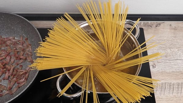 Dry spaghetti being added to a pot of boiling water beside a pan of pancetta